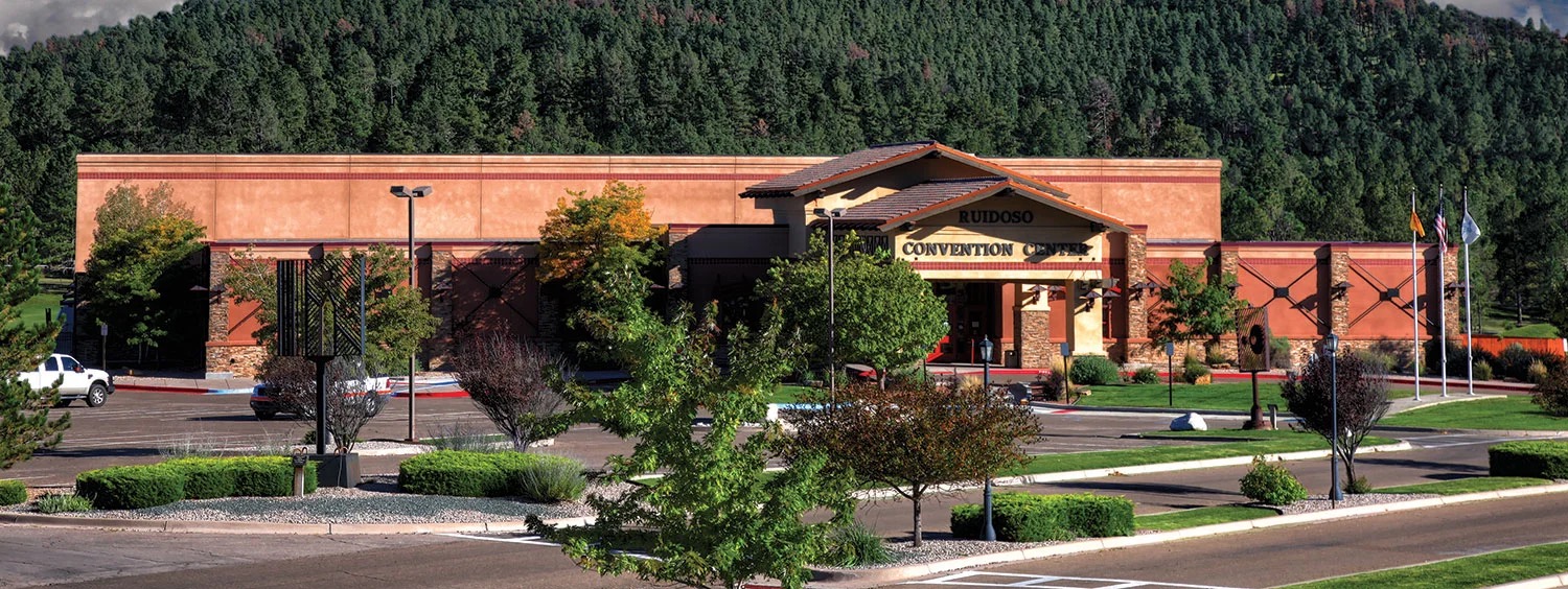 The Convention Center’s front entrance and portico by day, terracotta stucco walls, a red tile accent band, and forested mountains behind the building.